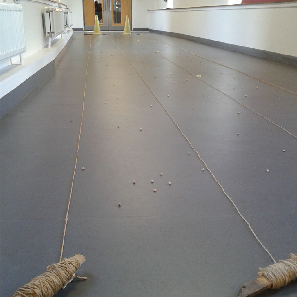 Grey corridor floor with yellow cones at one end and four lengths of string stretching towards the camera, evenly spaced across the corridor. There are random small pebbles scattered between the strings