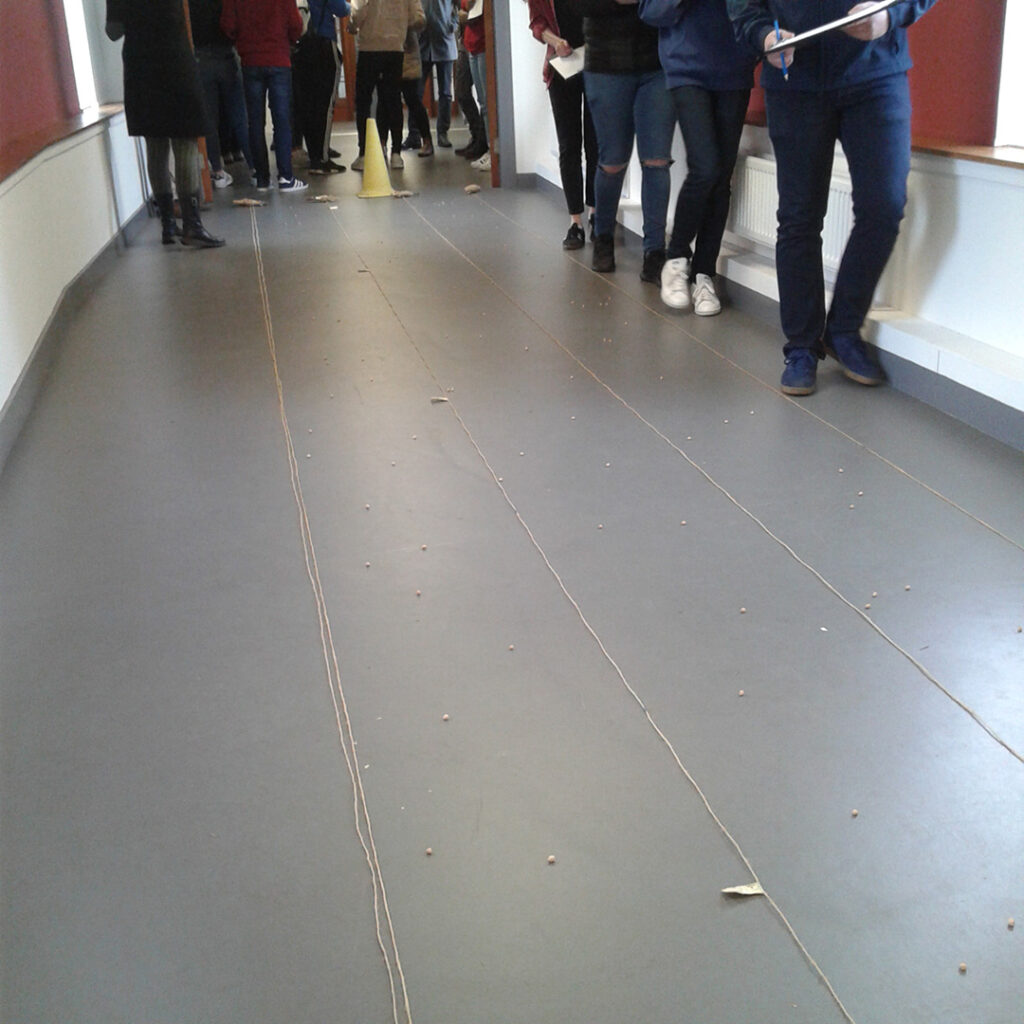 Grey corridor floor with yellow cones at far end and people standing around the cones.  FFour lengths of string stretch towards the camera, evenly spaced across the corridor. There are random small pebbles scattered between the strings, and four people walking in single file down one side of the corridor
